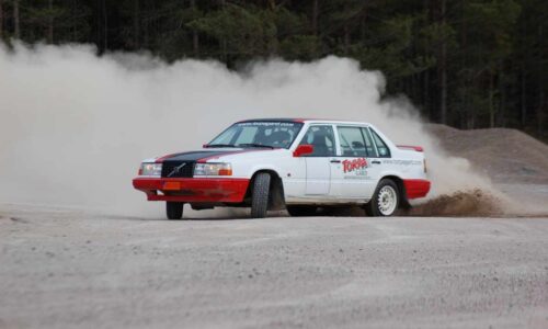 En orange-skyltad rallybil kör på tvären, det dammar ordentligt runt om.