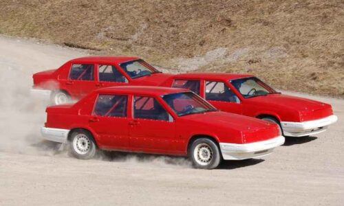 Tre röda folkracebilar kör nästan jämsides på en grusbana, det dammar lite runt hjulen.