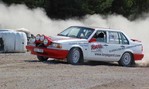 En vit/röd rallybil med ett stort damm-moln bakom. På sidan av bilen finns en stor logga och text.