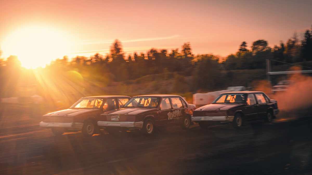 Två röda folkrace-bilar sedda snett från ovan. De kör efter varandra och ett damm-moln syns runt dem.