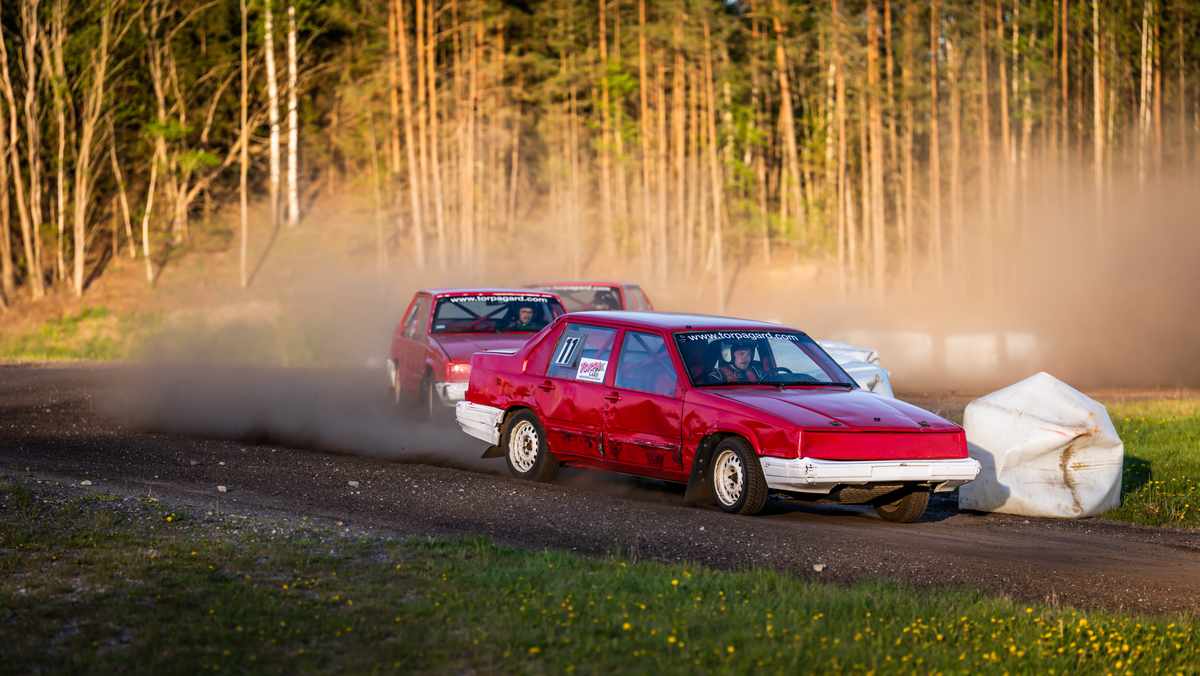 Två röda folkrace-bilar sedda snett från ovan. De kör efter varandra och ett damm-moln syns runt dem.